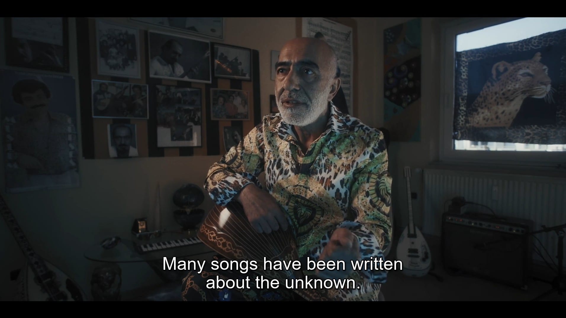 screenshot of orhan amuroglu in "aşk, mark ve ölüm" (cem kaya, 2022) who has a stringed instrument on his lap. he sits in a dark-lit room, behind him photographs and a leopard image. he also wears a shirt with leopard pattern. he says, "many songs have been written about the unknown."