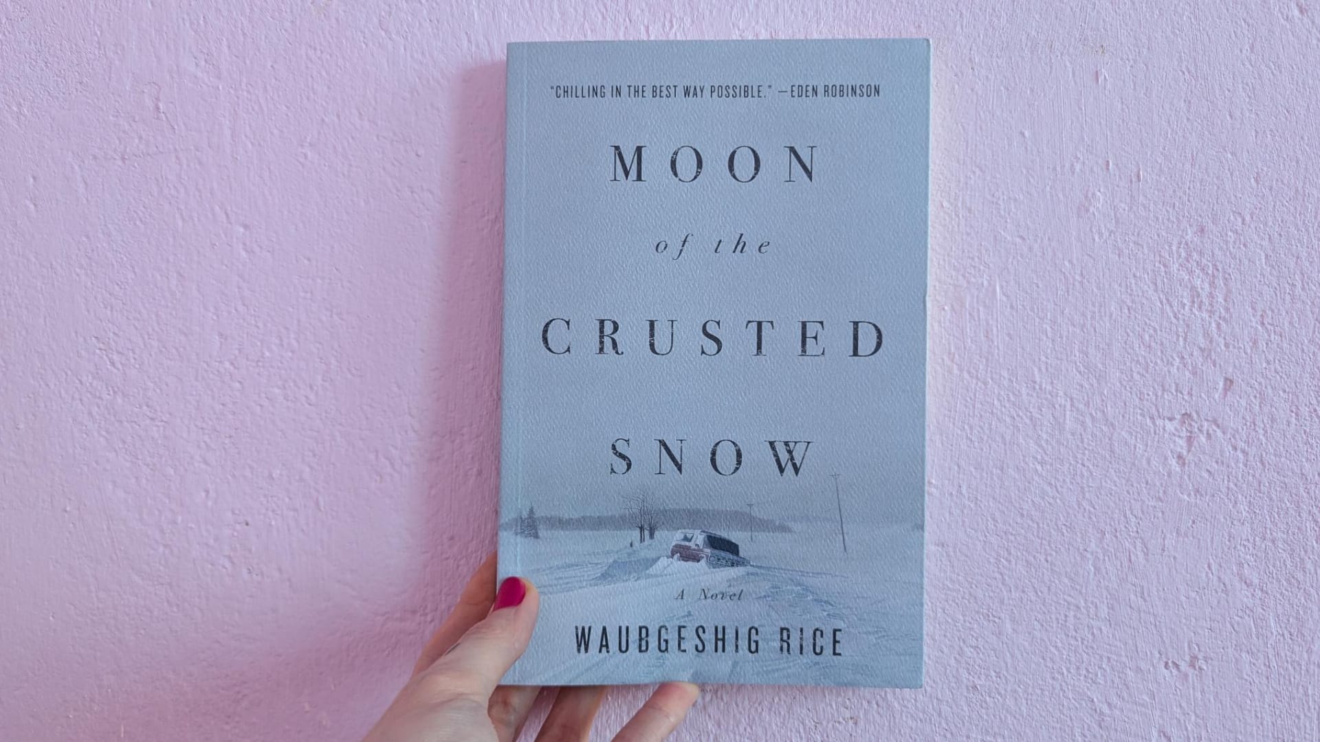 photo of my hand holding a copy of "moon of the crusted snow" by waubgeshig rice in front of a pastel pink wall.
