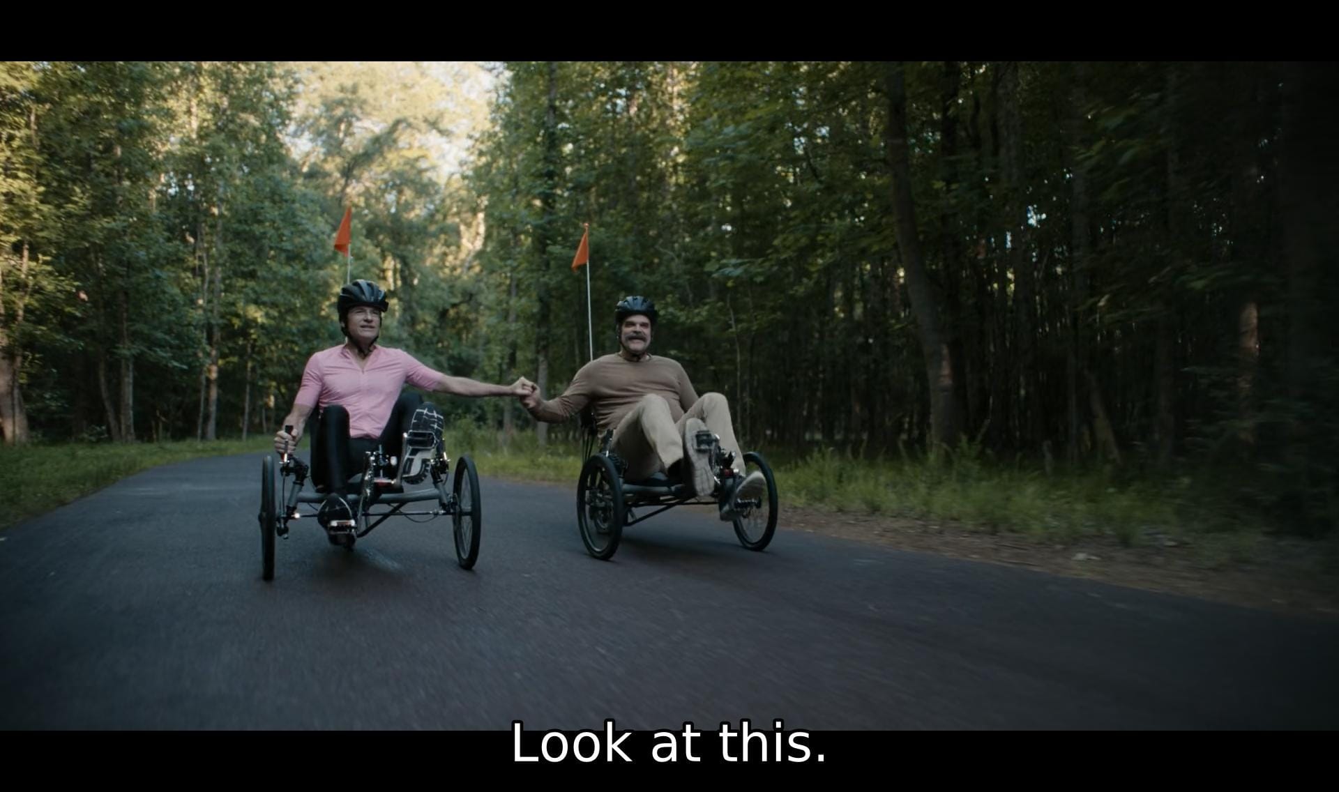 screenshot of dtf st. louis (s01e04) featuring clark and floyd riding their bikes in the woods. clark says, "look at this." they're holding hands.